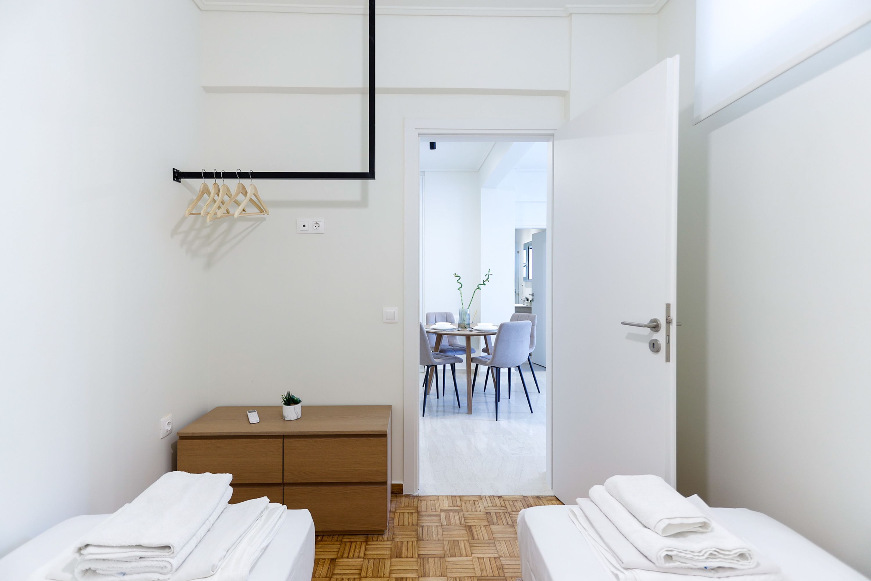 A modern hotel room with two beds, a wooden dresser, a small table with a plant, and a view into the dining area.