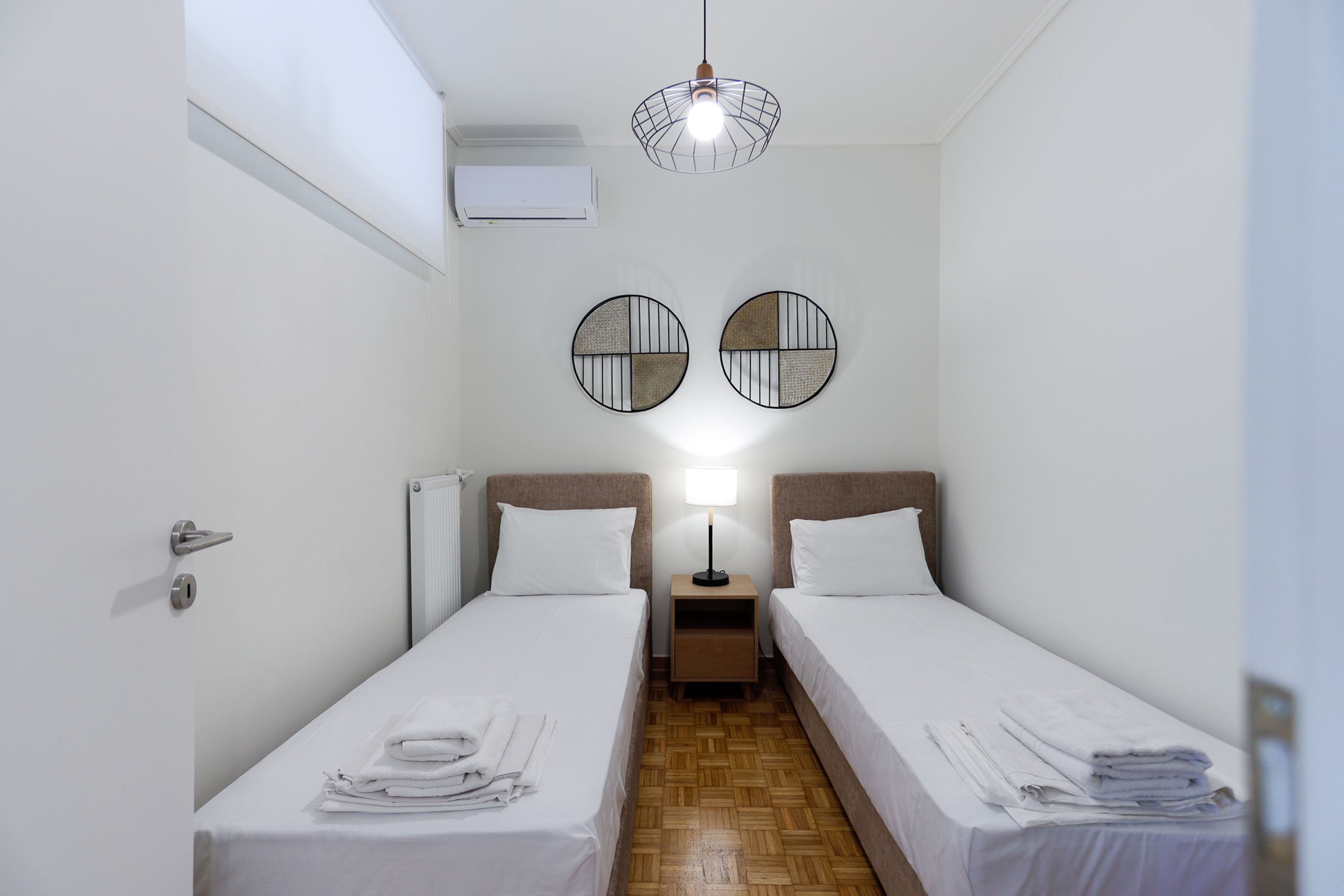 A hotel room with two single beds, a wooden floor, a wall-mounted air conditioner, and a ceiling light fixture.