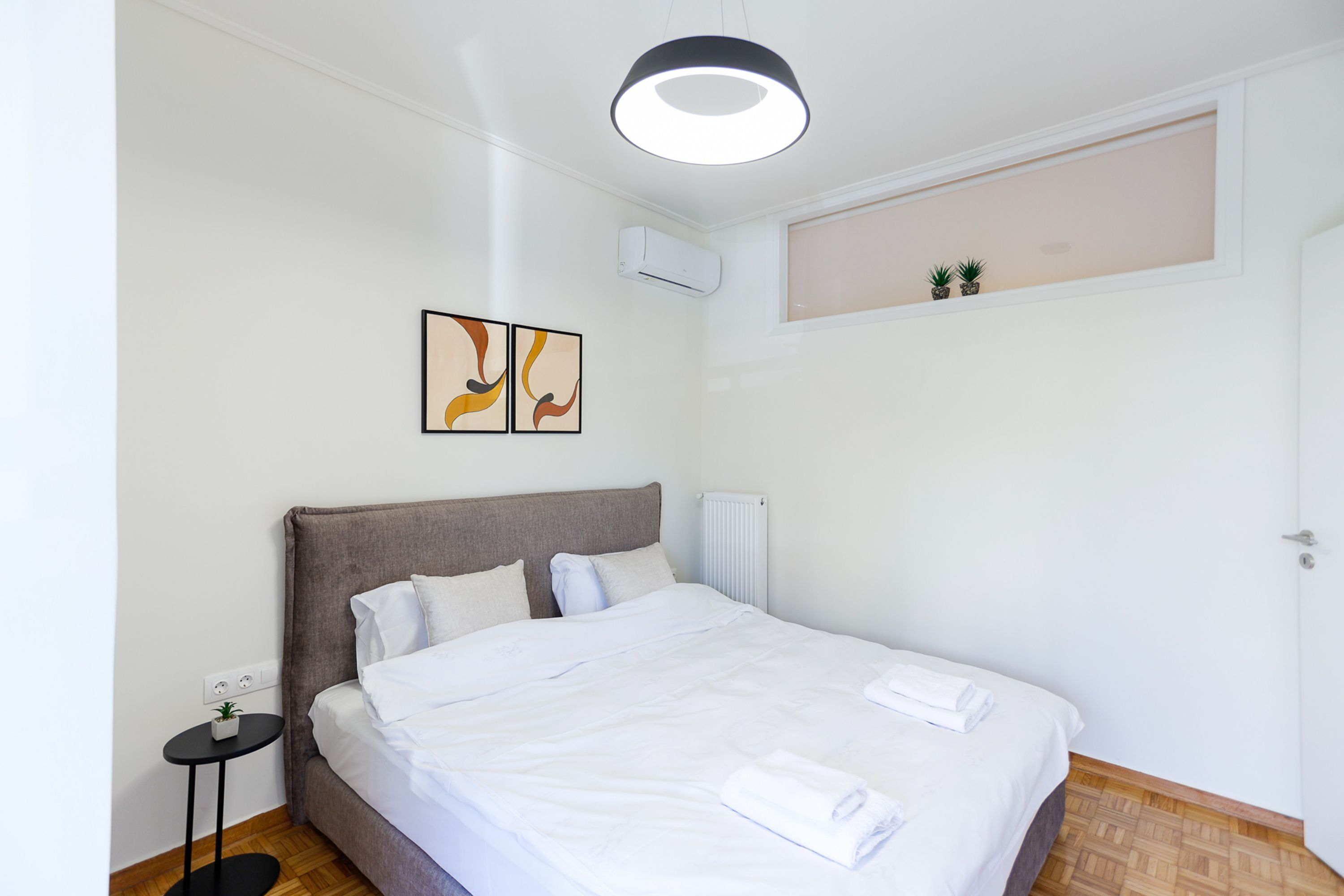 En pænt redt seng med hvide sengetøj, et natbord med en plante og en vægmonteret aircondition.