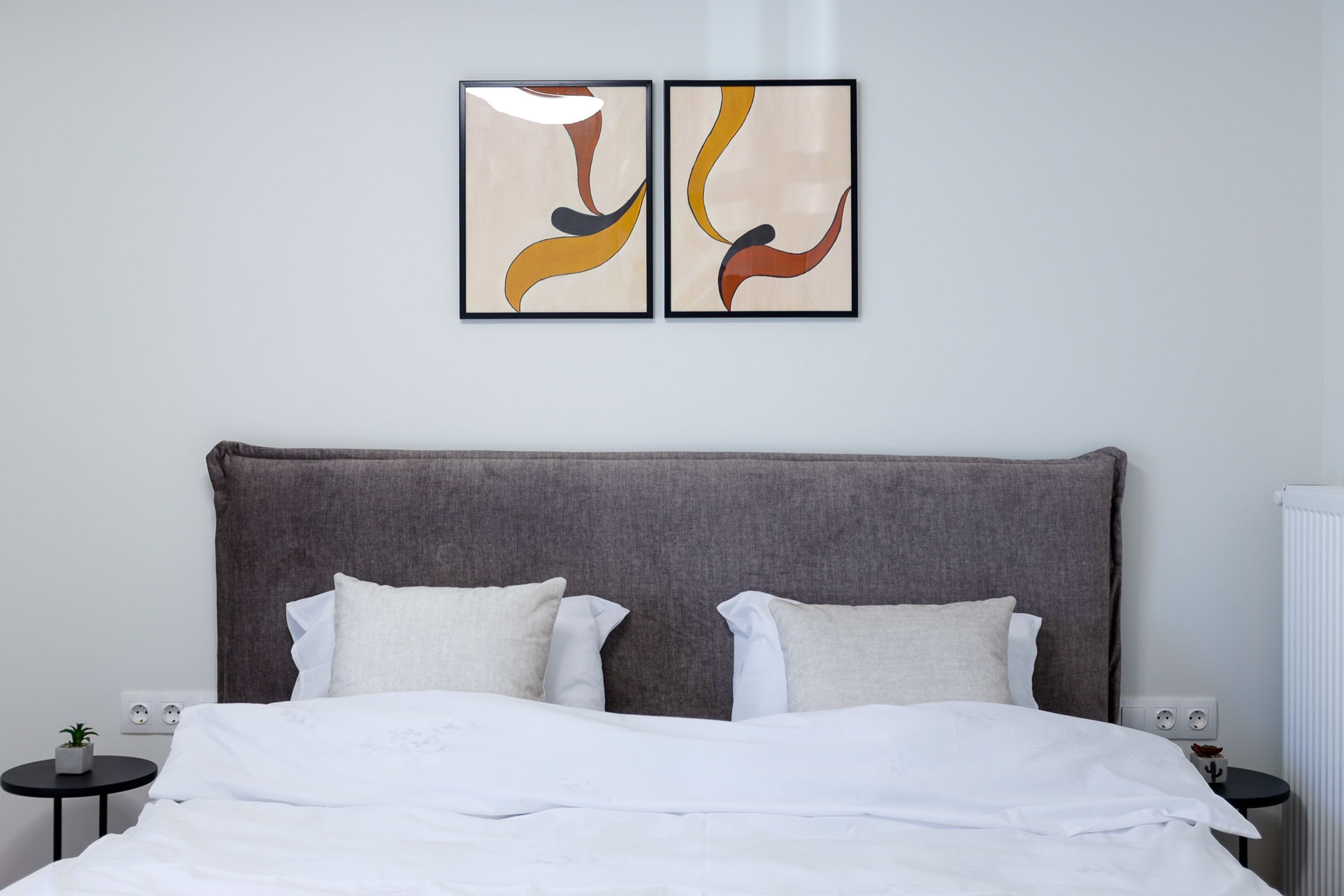 Un lit bien fait, avec du linge de lit blanc et deux oreillers, une tête de lit rembourrée de couleur grise et deux tableaux abstraits accrochés au mur.