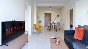 A modern living room with a flat-screen TV, a sofa, a coffee table, and a dining area with chairs and a table. - Modern 2-bedroom Apartment in Downtown Glyfada (Glyfada)