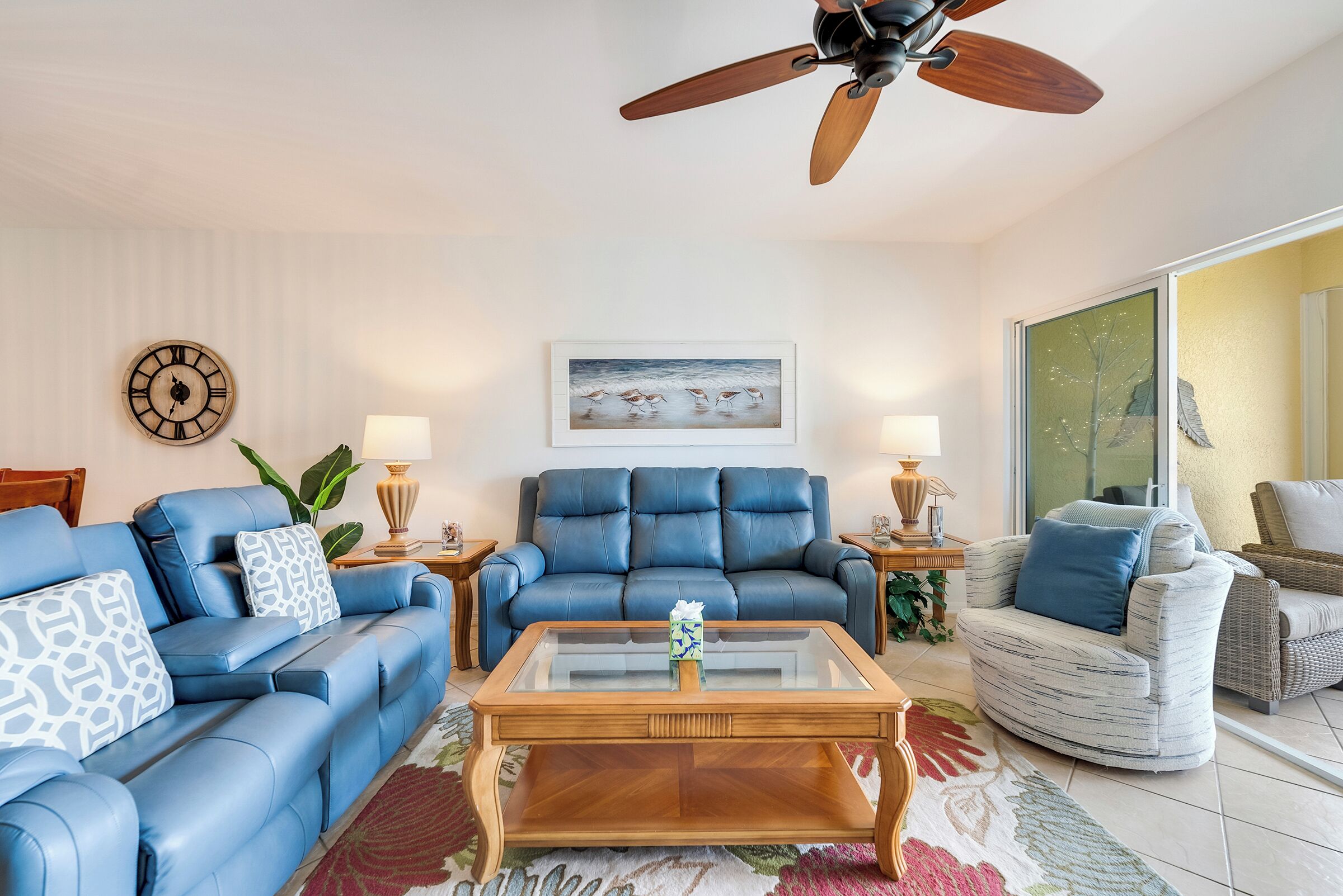 Ein Wohnzimmer mit blauem Sofa, Ledersessel, Glastisch, Holzboden und Deckenventilator.