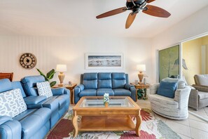 Ein Wohnzimmer mit blauem Sofa, Ledersessel, Glastisch, Holzboden und Deckenventilator.