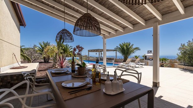 An outdoor dining area with a view of the pool and ocean, featuring a table set with plates, cutlery, and bottles.