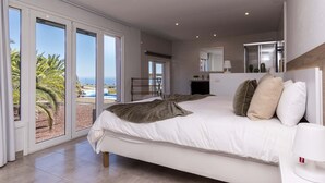 A spacious bedroom with a large bed, a desk, and a view of the beach through floor-to-ceiling windows.