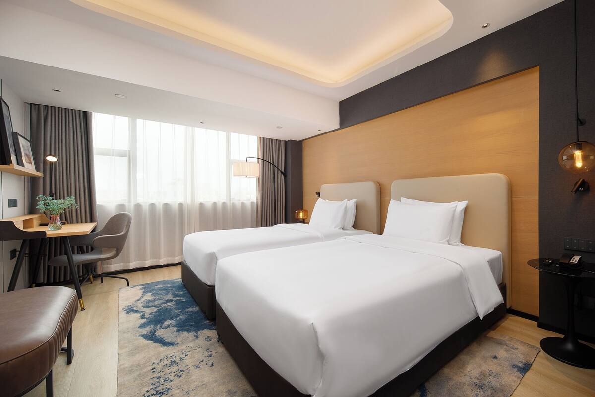 A hotel room with two beds, a desk with a chair, a small table with a phone, and a large window with curtains.
