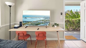 Une pièce comprenant un bureau, deux chaises, un grand tableau encadré représentant un paysage urbain côtier, une lampe et une porte donnant sur un jardin.