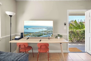 Una habitación con escritorio, dos sillas, un cuadro grande enmarco con una vista urbana costera, una lámpara y una puerta que da a un jardín.