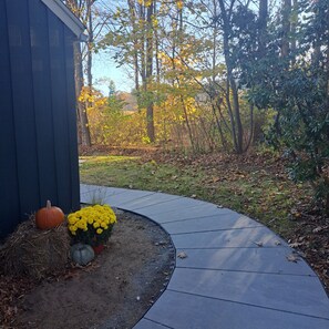 Un chemin sinueux mène à un bâtiment aux couleurs sombres, entouré d’arbres aux feuilles d’automne et d’un petit jardin où poussent des fleurs jaunes et des courges.