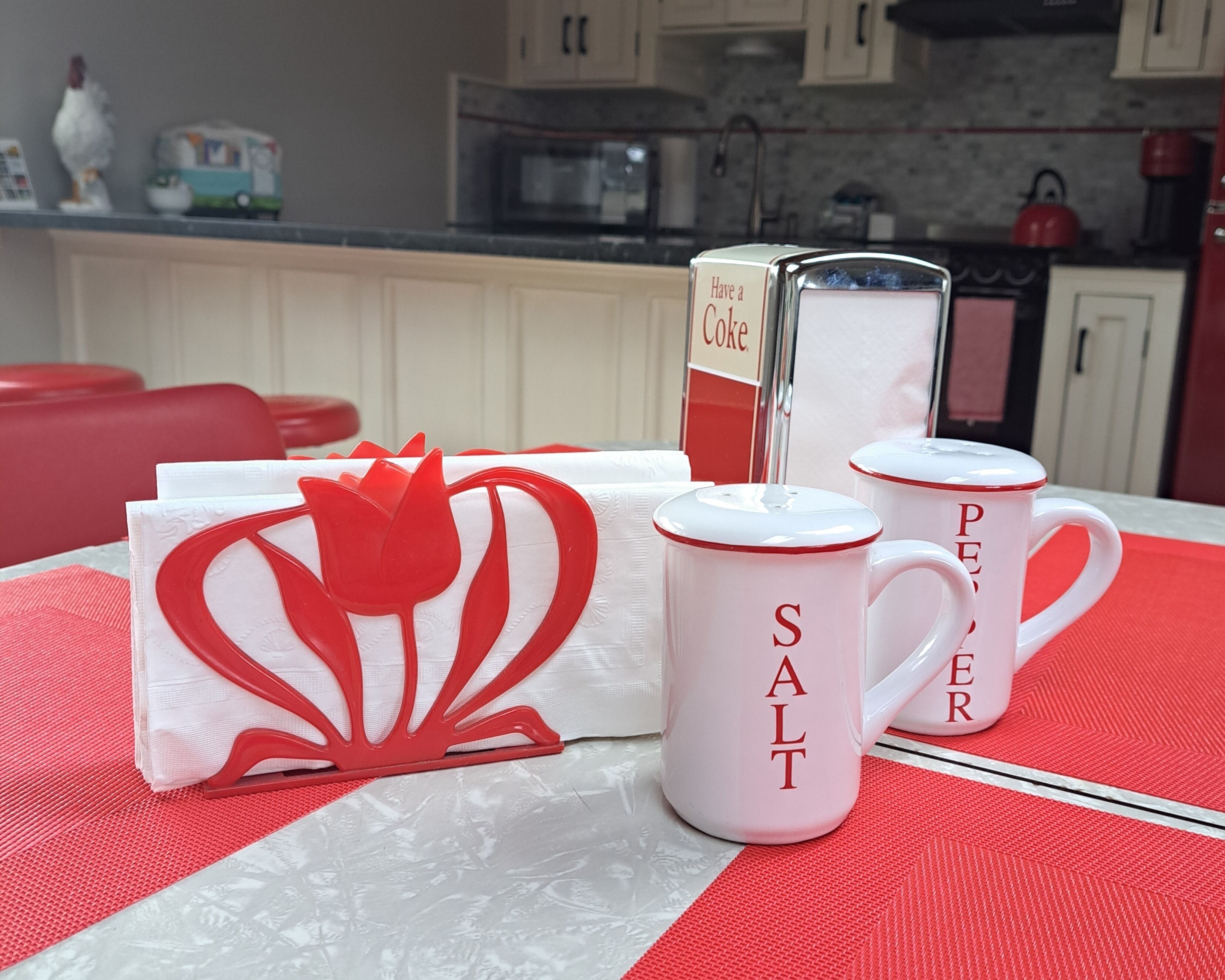 A kitchen with white cabinets, a microwave, and a red kettle. There are two white mugs with red text and a red and white decorative item on the table.