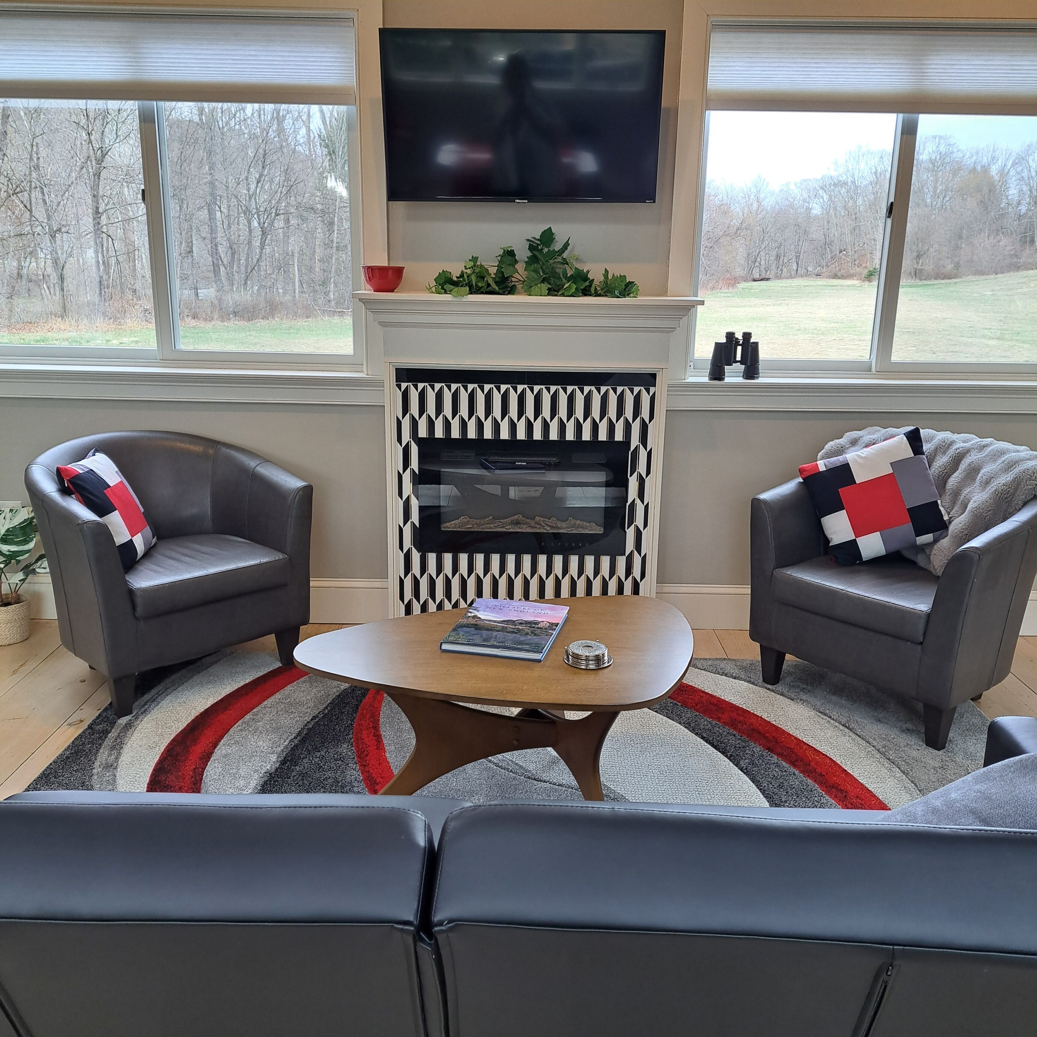 A living room with two grey armchairs, a round coffee table, a fireplace with a black and white patterned surround, and a TV mounted above the fireplace.