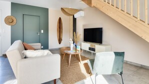 Een moderne woonkamer met een bank, salontafel en een televisie. Er is een houten trap en een deur, waar een hoed aan hangt.