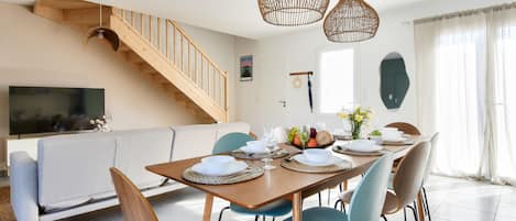 A dining area with a wooden table set for four, surrounded by chairs, a staircase, and a television in the background.