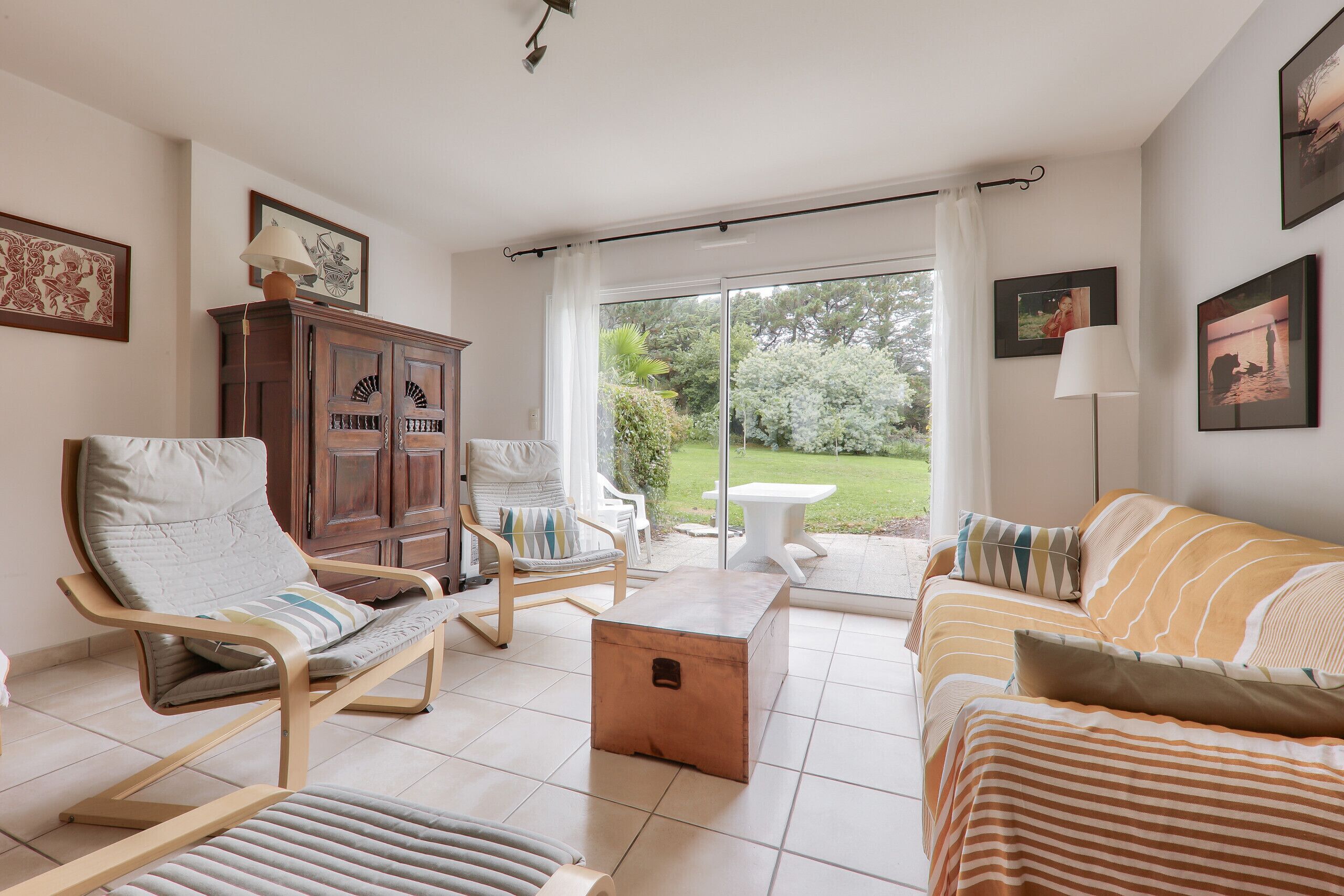 Ein Wohnzimmer mit einer Couch, Sesseln, einem Couchtisch und einem Schrank. Ein Glastür ermöglicht den Zugang zu einem Garten.