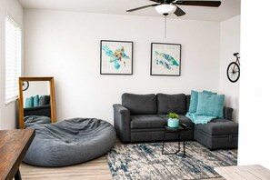 Uma sala de estar com sofá cinza, um bean bag, uma mesa de centro, um ventilador de teto e duas obras de arte emolduradas.