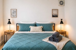 A neatly made bed with teal bedding, two bedside lamps, and a wall-mounted air conditioner.