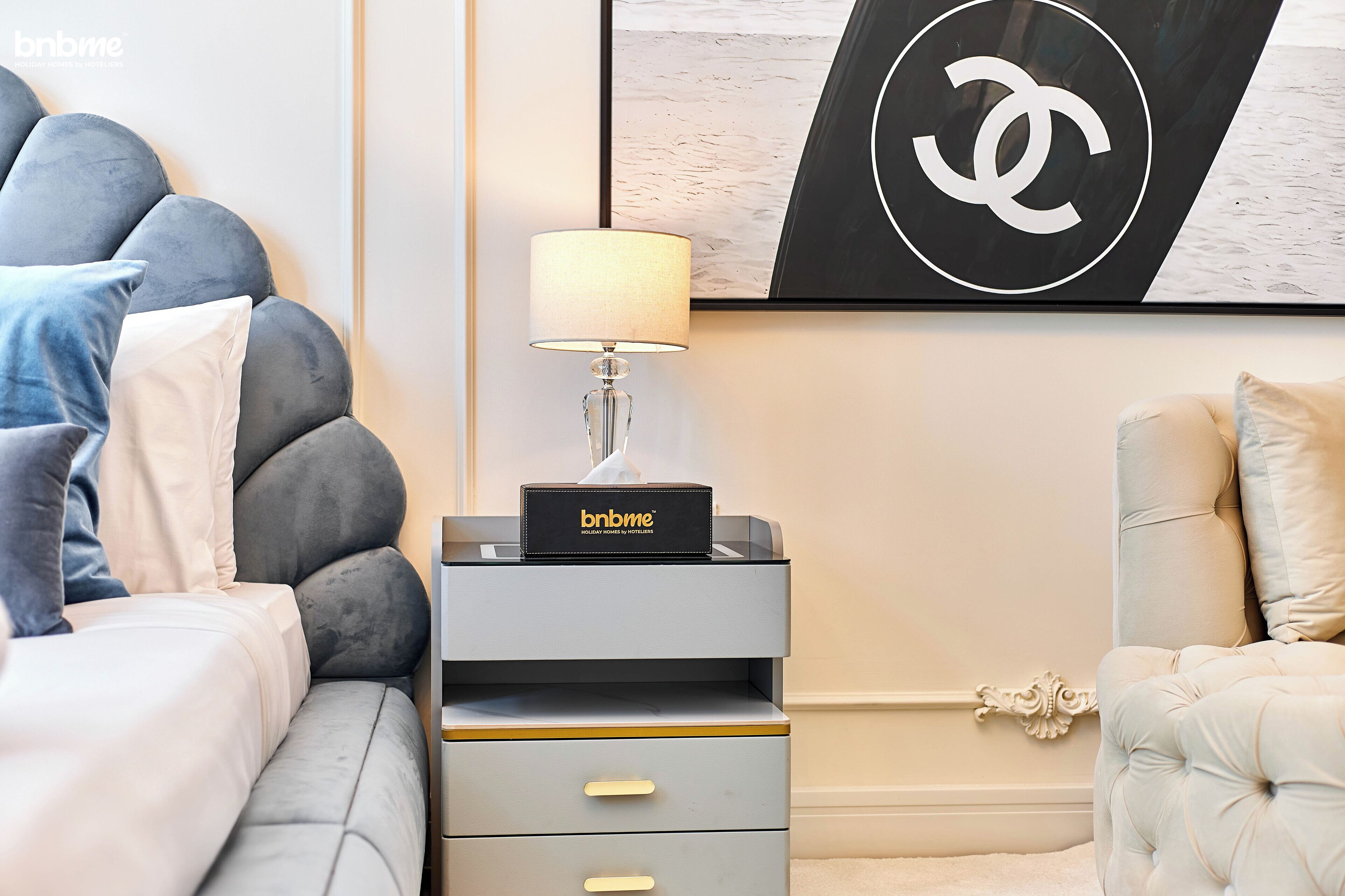 Une chambre à coucher bien rangée, avec un lit, une table de chevet dotée d’une lampe, un logo encadré accroché au mur et un élément décoratif mural.