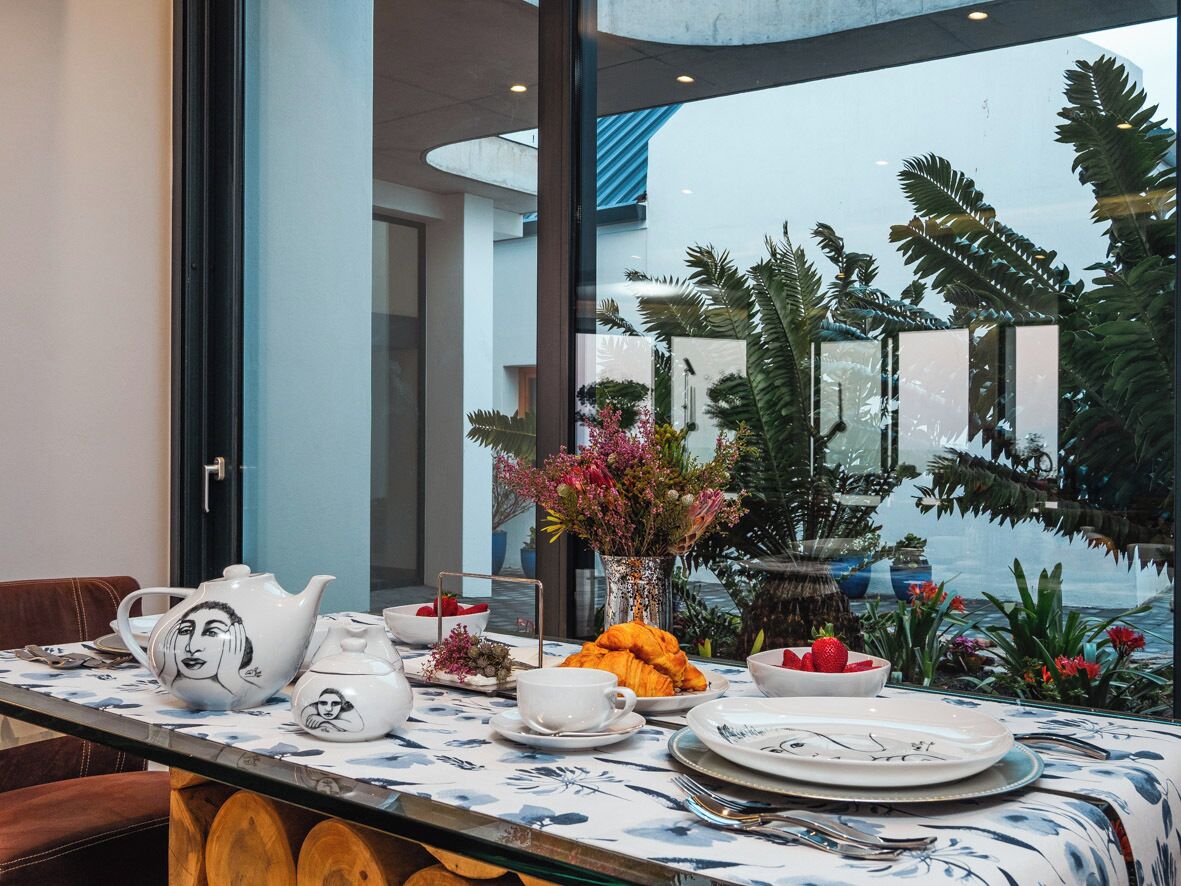 Een eettafel gedekt met borden, kopjes, een theepot en een schaal fruit, met uitzicht op een tuin en palmbomen door een raam.