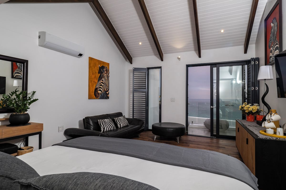 A modern bedroom with a large bed, a black leather sofa, a small round ottoman, a wooden side table with a plant, and a view of the sea through a glass door.