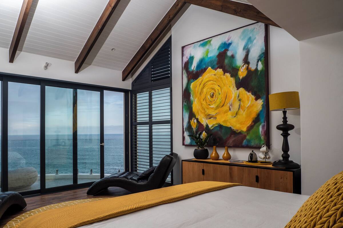 A modern bedroom with a large window, a bed, a painting of a yellow rose, and a nightstand with a lamp.