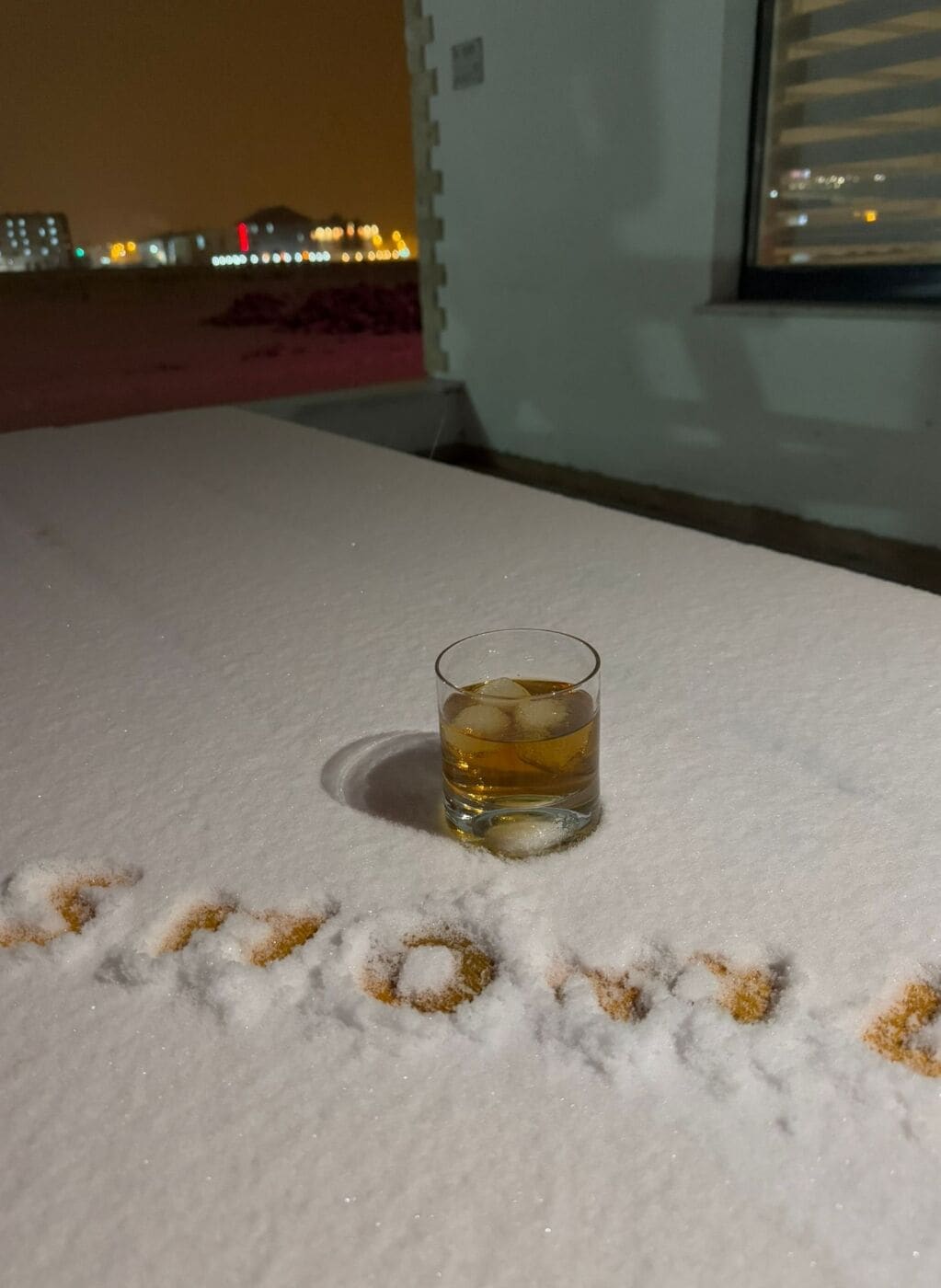 A glass of amber liquid on a table with snow footprints spelling "LOVE" in the snow.