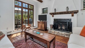 A cozy living room with a fireplace, wooden beams, a stone fireplace surround, a television on a wooden cabinet, and a large window with a view of the outdoors. - Wolf Creek House (Black Mountain)