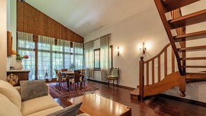 A spacious living room with a wooden staircase, a dining area with a table and chairs, and a sofa with cushions.