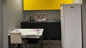 Een kleine keuken met gele kastjes, een witte koelkast, en een eethoek met een tafel en stoelen.
