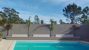 A rectangular swimming pool with a clear blue water surface, surrounded by a low grey wall with steps leading up to it. There are palm trees planted in the wall, and a clear sky above. - Viana do castelo Villa rioneiva (Alvarães)