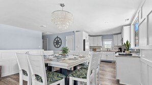 A modern kitchen with white cabinetry, a dining area with white chairs and a chandelier, and a view of the city through large windows.