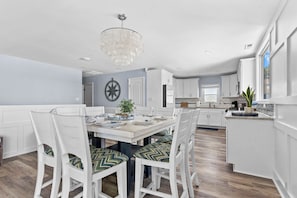 A modern kitchen with white cabinetry, a dining area with white chairs and a chandelier, and a view of the city through large windows.