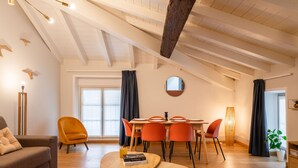 Amplia sala de estar con techo alto, vigas de madera, una mesa de comedor con sillas color naranja, un sofá y una mesita con libros.