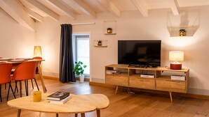 Una sala moderna con un mueble de televisión de madera, un televisor de pantalla plana, una zona de comedor con sillas color naranja y una mesita con un libro y un vaso.