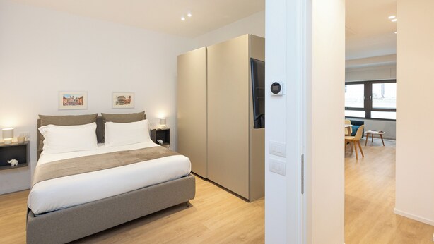 Une chambre à coucher moderne avec un grand lit, des tables de chevet, une armoire et une vue sur le salon.