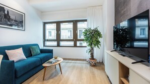 Un salon avec un canapé bleu, une table basse, une plante en pot, un téléviseur à écran plat et de grandes fenêtres donnant sur la ville.