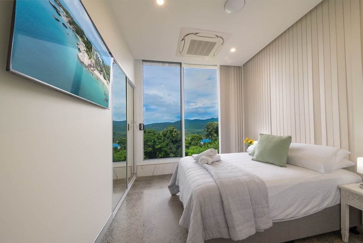 A bedroom with a large bed, a nightstand, a window with a view of greenery, and a ceiling vent.