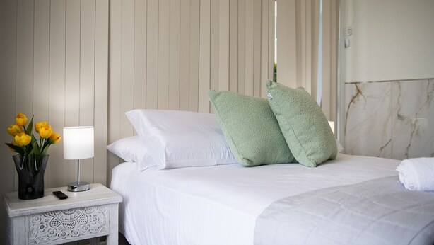 Habitación de hotel con cama, ropa de cama blanca, mesita de noche con lámpara y un jarrón de flores, y pared de mármol.