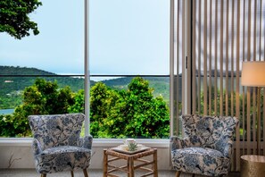 Ein Zimmer mit zwei gemusterten Sesseln, einem kleinen Tisch und Blick auf eine grüne Landschaft durch große Fenster.