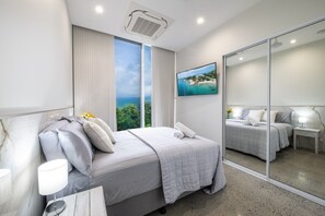 Ein modernes Hotelzimmer mit einem großen Bett, einem Nachttisch mit Lampe, Meerblick und einem großen Spiegel.
