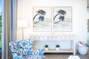 A room with a blue patterned armchair, a white console table with drawers, and two wall-mounted artworks depicting palm trees.