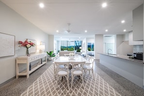 A modern kitchen with a dining area, featuring a central island, white cabinetry, and a view of the outdoors. - Airlie View - Coastal Escape with Sea Views (Airlie Beach)
