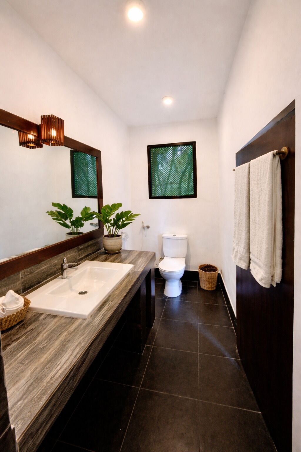 A modern bathroom with a wooden vanity, a white toilet, and two potted plants.