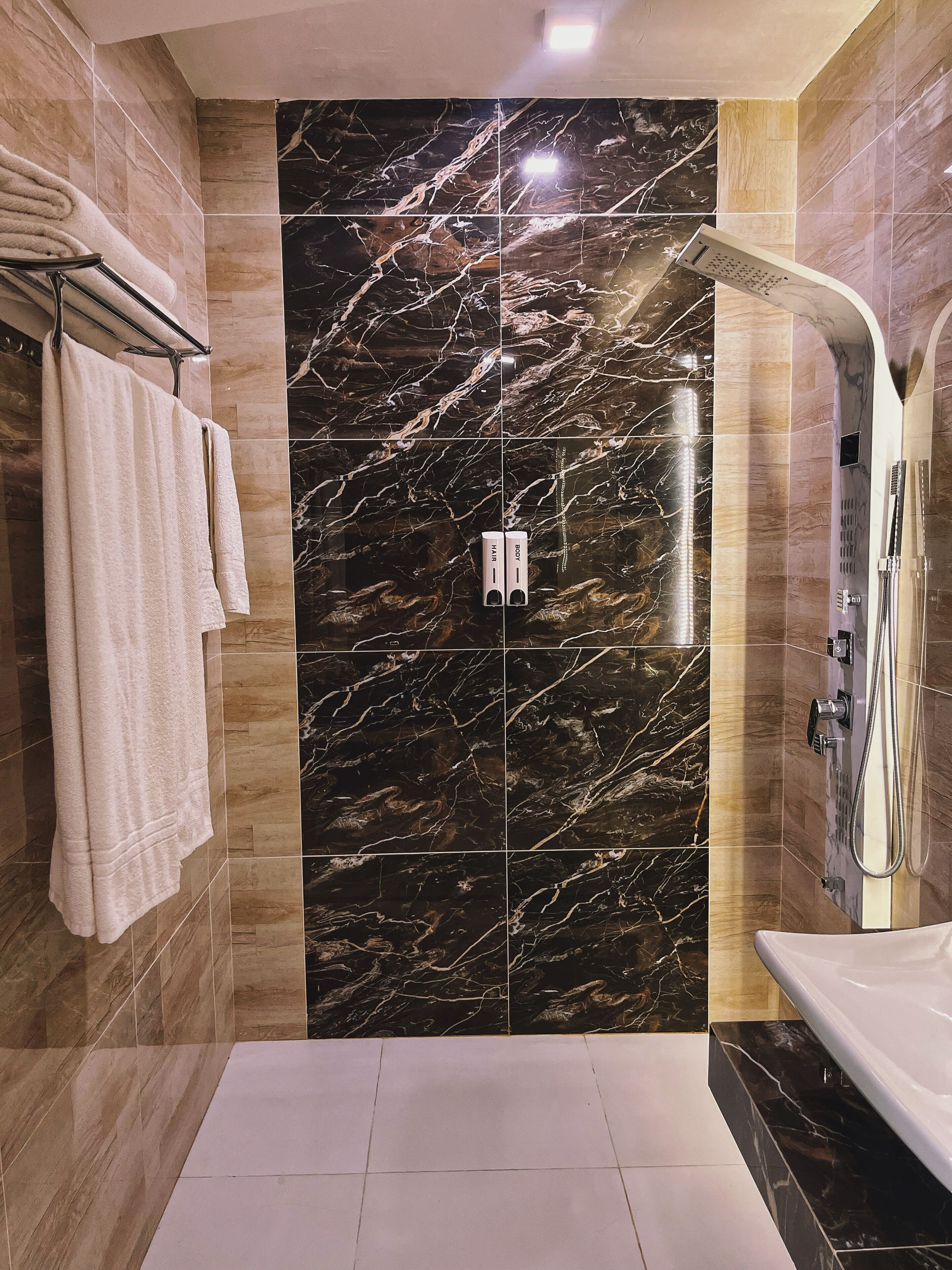 A modern bathroom with a marble wall, a shower with a handheld showerhead, and two towels on a rack.