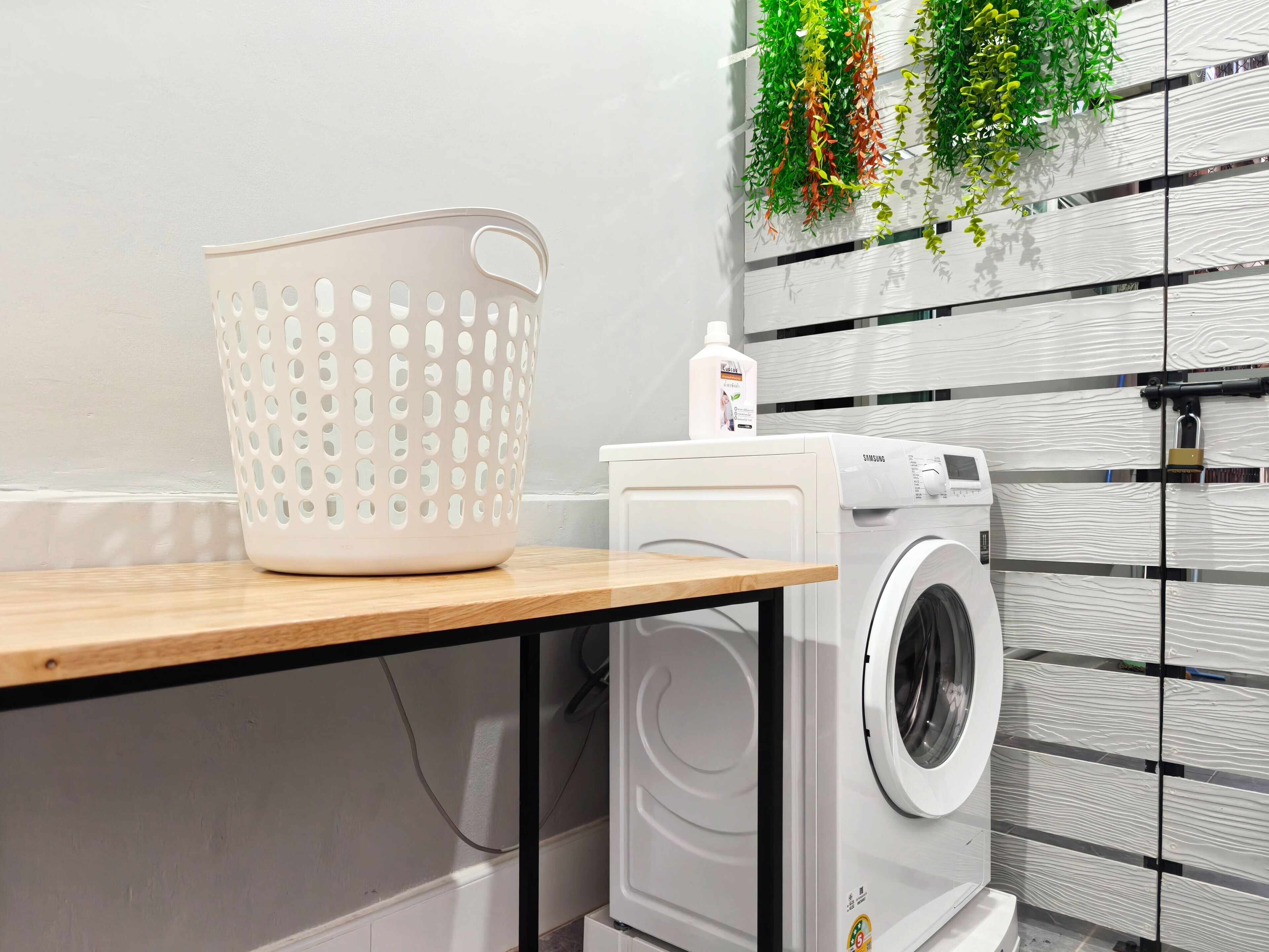 A laundry area with a washing machine, a white laundry basket, and a bottle of detergent on a wooden table.