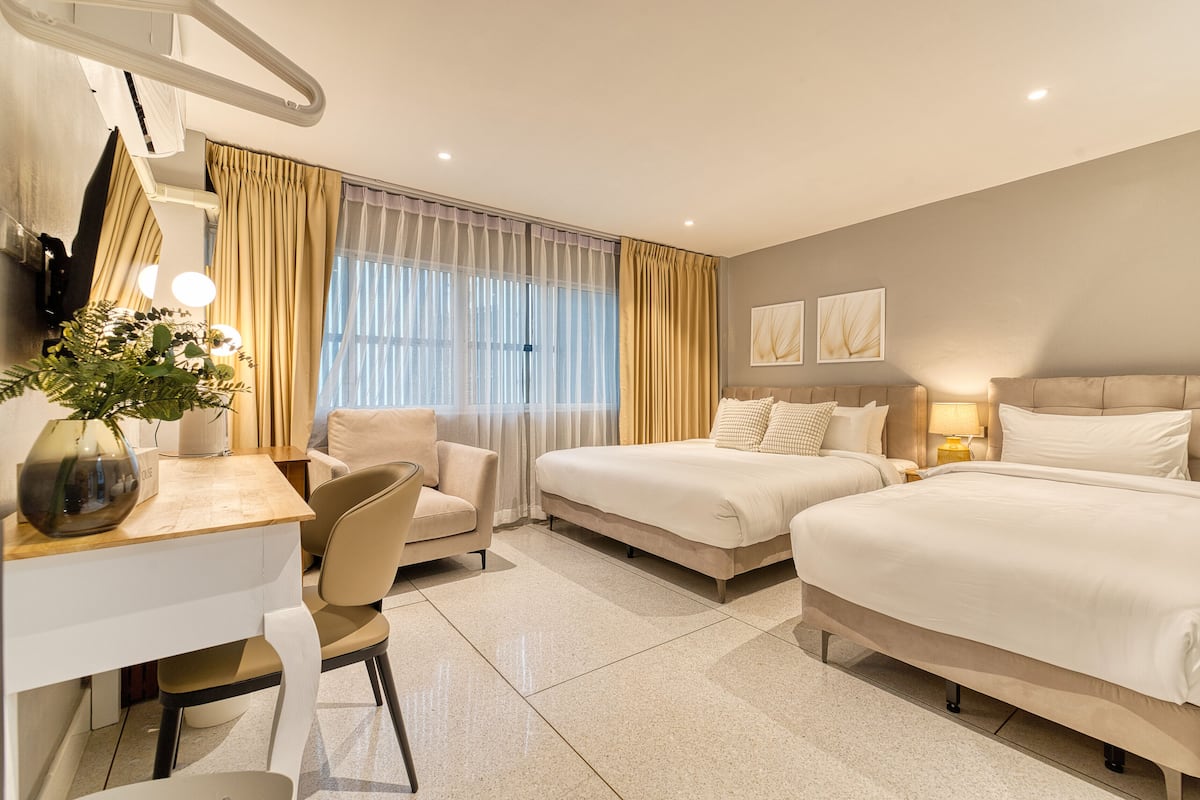Ein Hotelzimmer mit zwei Betten, einem Schreibtisch mit Stuhl, einer Topfpflanze und einem Fenster mit Vorhängen.