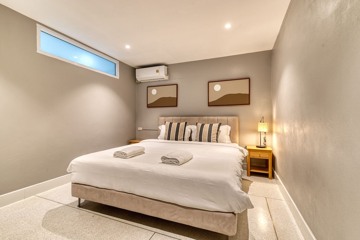 Ein modernes Schlafzimmer mit einem großen Bett, einem Oberlicht, einer Klimaanlage, einem Nachttisch mit Lampe und zwei gerahmten Bildern an der Wand.