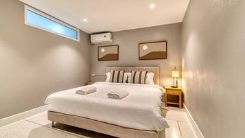 A modern bedroom with a large bed, a skylight, an air conditioning unit, a nightstand with a lamp, and two framed pictures on the wall.
