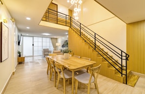 Amplia sala de estar con una mesa de comedor de madera, asientos cómodos, una escalera con barandilla negra y un ventanal que proporciona luz natural.
