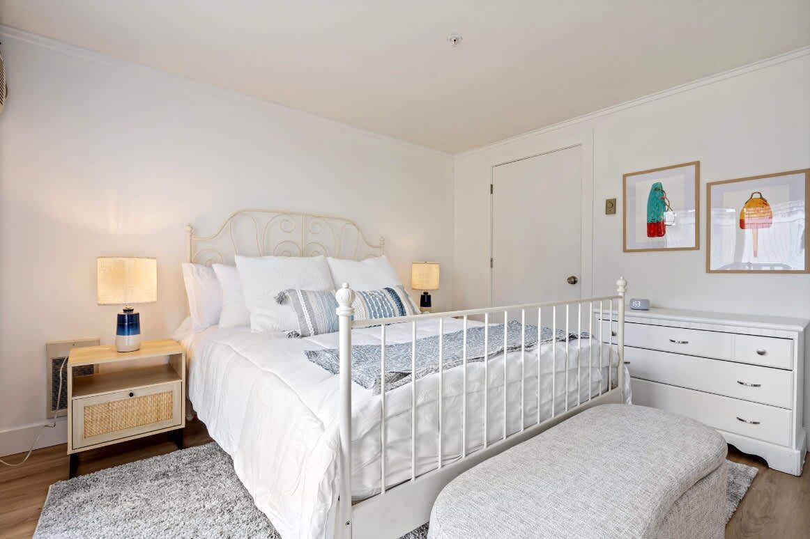 A bedroom with a white bed, two bedside tables with lamps, a white dresser, and a door.