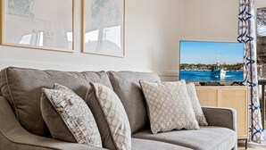 Een grijze bank met gekleurde kussens, een flatscreen televisie toont een kustlandschap, en er hangt een lijst met kunstwerken aan de muur.